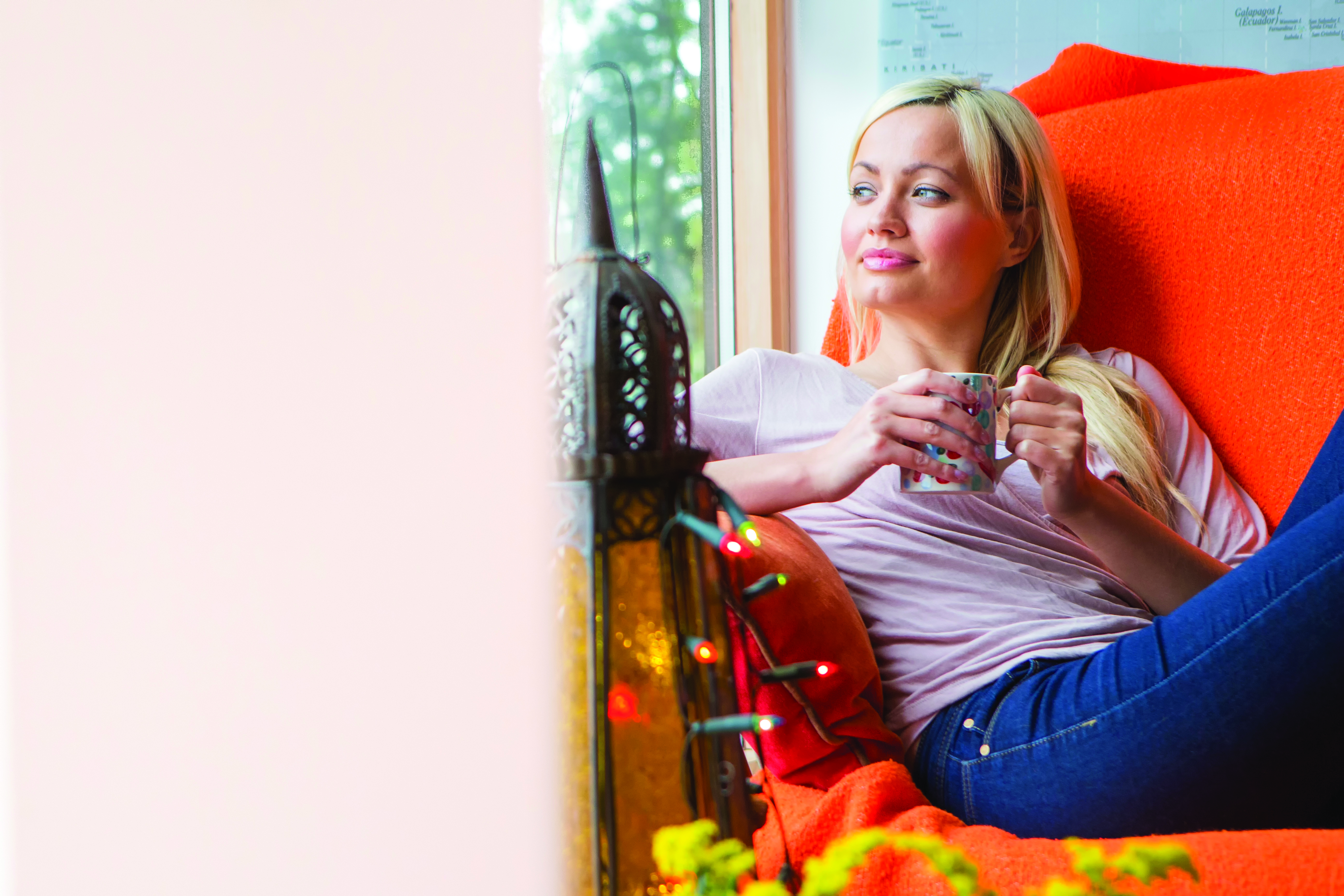 A happy blonde smiles as she sits on her sofa with a hot drink and looks out of the window. There are small decorative lights in front of her and large copy space in the direction that she is looking in.
